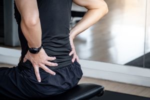 Sport man feeling lower back pain or spine pain while sitting on workout bench in fitness gym. Male athlete suffering from sport injuries symptoms.